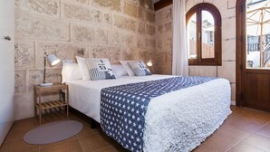 A bedroom with a stone wall, a wooden ceiling, a bed with a white and blue patterned bedspread, two bedside lamps, and a window with a view of buildings. - Can Xisco (Sineu)