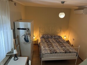 A bedroom with a bed, bedside lamps, a wardrobe, a desk with a lamp, and a hanging bag.