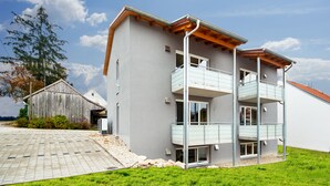 A modern multi-story building with balconies, a paved driveway, and a wooden barn in the background. - 5 large apartments, for a maximum of 4 people in an idyllic location near Munich (Billingsdorf)