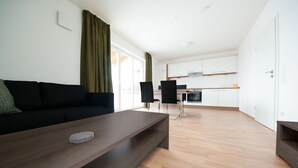 A modern living room with a dark brown coffee table, a black sofa, and a dining area with two black chairs. A kitchen with white cabinets and a wooden countertop is visible in the background. - 5 large apartments, for a maximum of 4 people in an idyllic location near Munich (Billingsdorf)