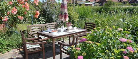 A patio with a table set for two, surrounded by chairs, in a garden with flowering plants and a view of houses in the background.