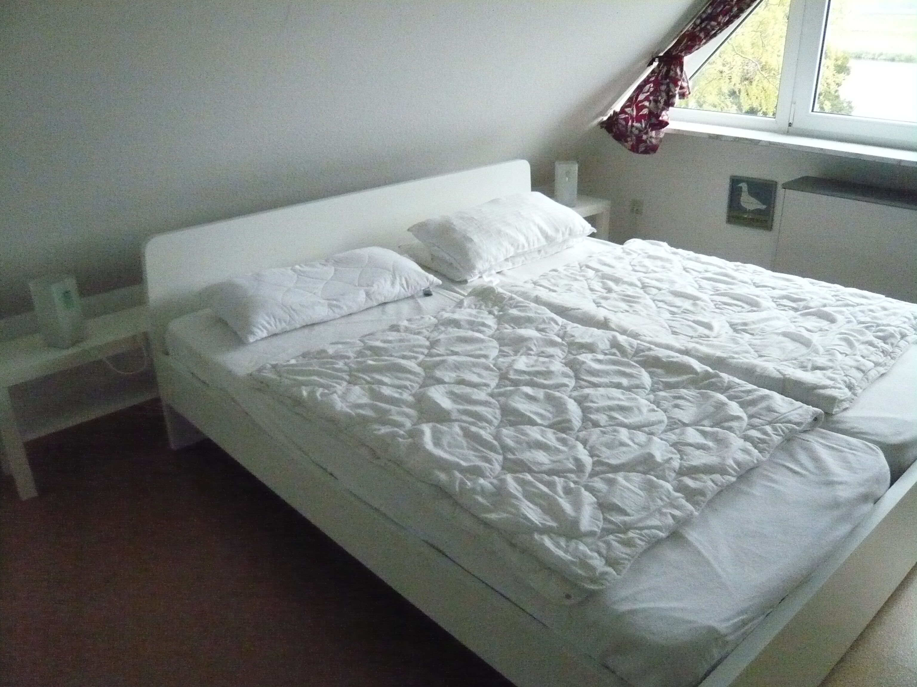 Una camera semplice con un letto singolo, un poggiatesta essenziale, un piccolo comodino e una finestra con tenda.