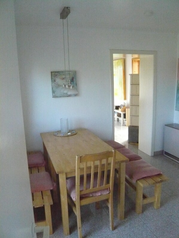 A dining area with a wooden table and benches, a hanging light fixture, and a view into another room.