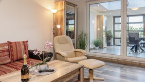 Ein modernes Wohnzimmer mit einem Holztisch, einem beigen Sessel und einer Couch mit gemusterten Kissen.