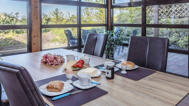 Ein Frühstückstisch gedeckt mit Eiern, Brot, Butter und Kaffee in einem Raum mit großen Fenstern und Stühlen.