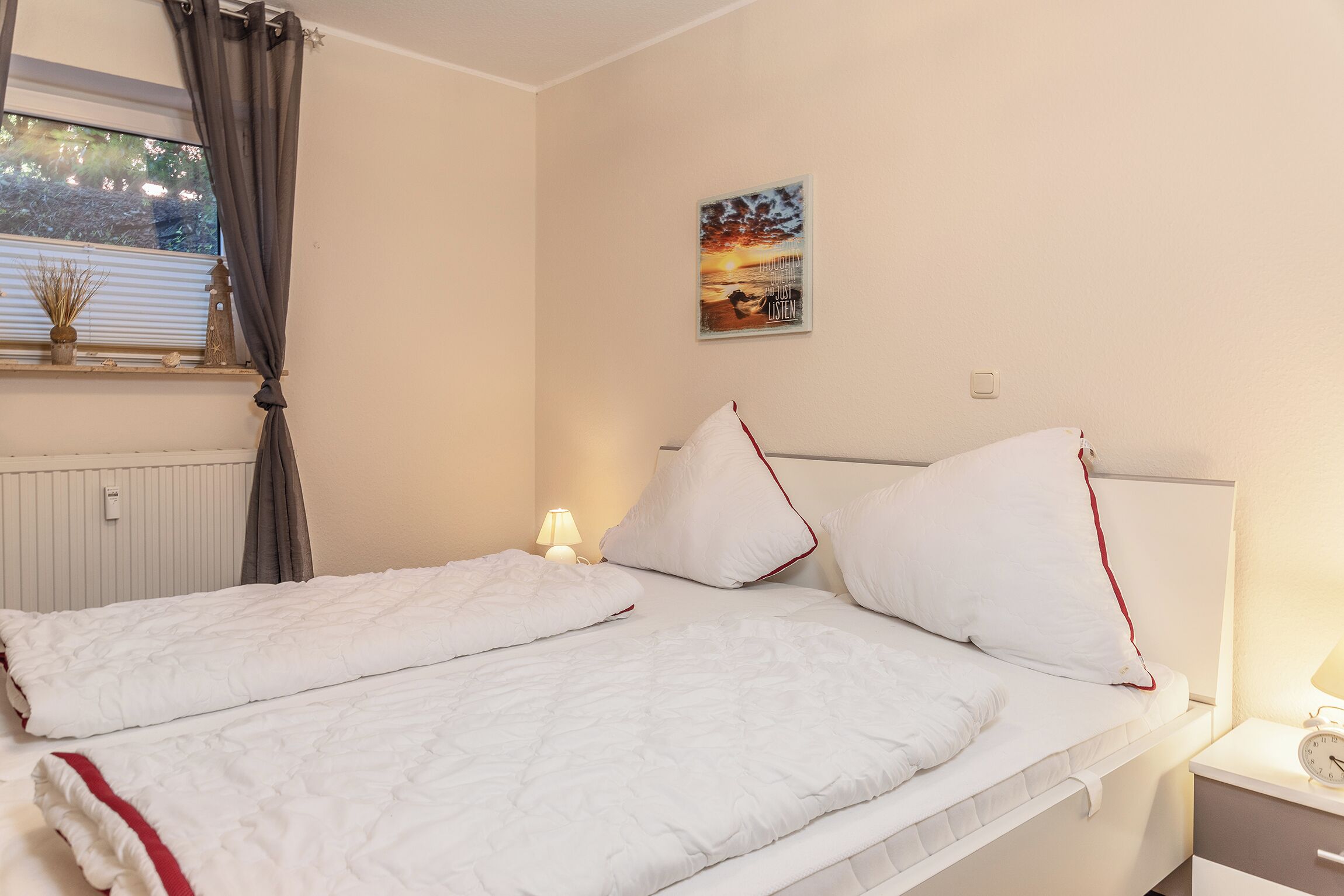 A neatly made bed with white bedding and red trim, a bedside lamp, and a wall clock.