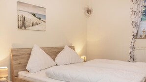 Ein ordentlich bezogenes Bett mit weißer Bettwäsche und Kissen, ein hölzerner Kopfteil, ein Nachttisch mit Lampe und ein Wandbild mit Strandmotiv.