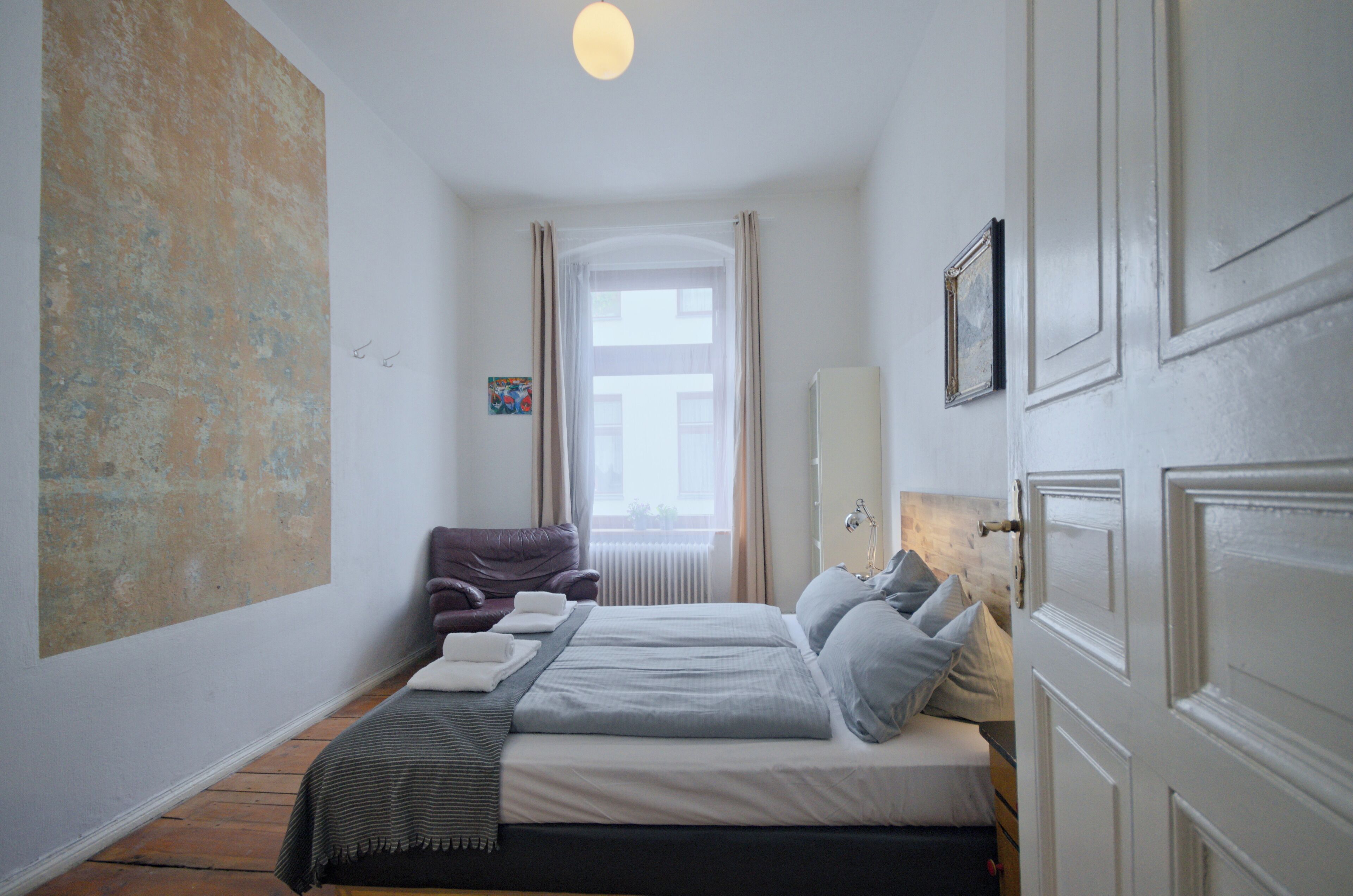 Una camera da letto con un grande quadro astratto, un letto con biancheria grigia, una poltrona viola e una finestra con tende.