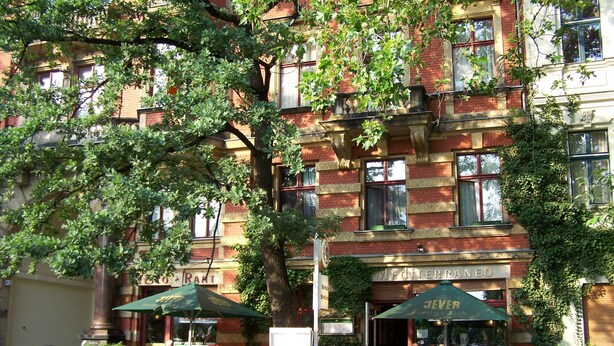 Tiilirakennuksessa on alakerrassa ravintola nimeltä ”Mediterraneo”. Ulkona on ulkoilualueita, joilla on vihreitä aurinkovarjoja.