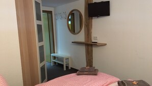 Ein Schlafzimmer mit einem Bett, einem kleinen weißen Sessel, einem Holzregal mit einem Fernseher und einem Spiegel an der Wand.