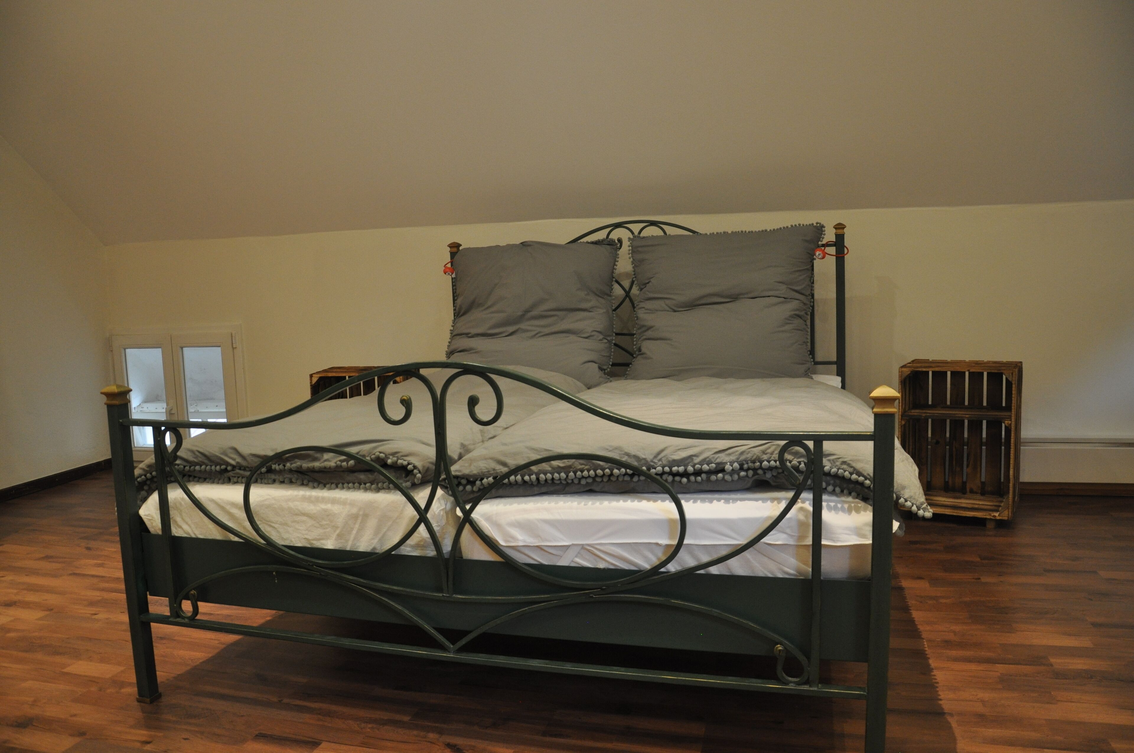 Een bed met metalen frame en grijze beddengoed in een kamer met houten vloer en een witte muur.