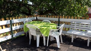 Un patio exterior con una mesa y sillas cubiertas por un mantel verde, rodeado por una cerca blanca y árboles.
