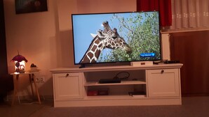 Un televisor sobre un mueble blanco con un control remoto y una mesita con una lámpara y una taza al lado.