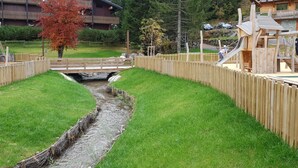 A wooden bridge over a small stream in a park area with playground equipment and a building in the background. - Châtel Portes du Soleil Free MultiPass 3-Star Apartment PDS Chairlifts 400 meters away (châtel)