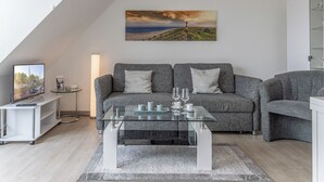 Een moderne woonkamer met een grijze zithoek, een glazen salontafel, een schilderij van een kustlandschap en een televisie op een meubel.'