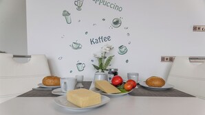 Een ontbijtje met een bord boter, kaas, tomaten en brood op een tafel tegen een witte muur met illustraties van koffie en thee.
