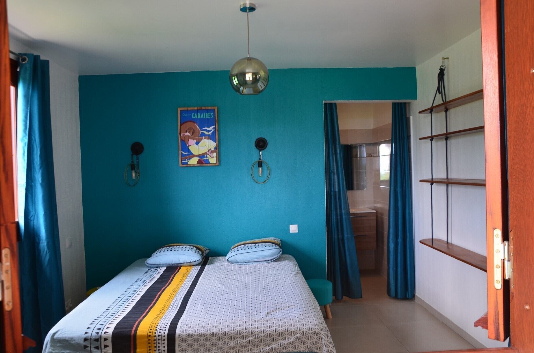 Ein Zimmer mit türkisfarbenen Wänden, einem Bett mit gemusterten Betttextilien, einer hängenden Leuchte und einer Tür zu einem anderen Raum.