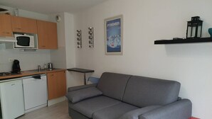 A compact kitchen with a microwave, coffee maker, and a small table. A grey sofa is positioned against the wall, and a poster is displayed above it.