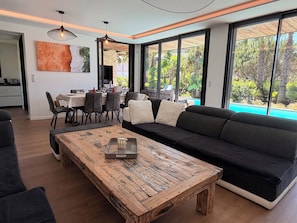 A modern living room with a wooden coffee table, black sofa, dining area with chairs, and large windows offering a view of a pool and greenery.