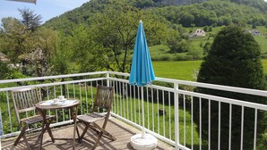 Ein Balkon mit Tisch und Stühlen, einem blauen Sonnenschirm und Blick auf ein grünes Tal und Berge.