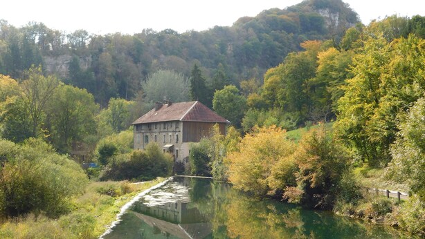 Ein Gebäude aus Stein mit einem Ziegeldach, umgeben von üppiger Vegetation und einem ruhigen Wasserlauf.