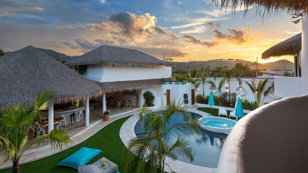 A resort with thatched-roof structures, a swimming pool, and a view of the sunset.