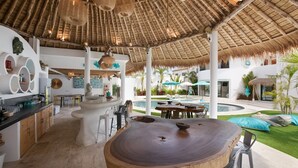 A spacious outdoor dining area with a thatched roof, a pool, and a bar area with stools and a counter.