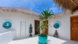 A serene courtyard with a central potted plant, a ladder, and statues against a white wall with thatched roofs.