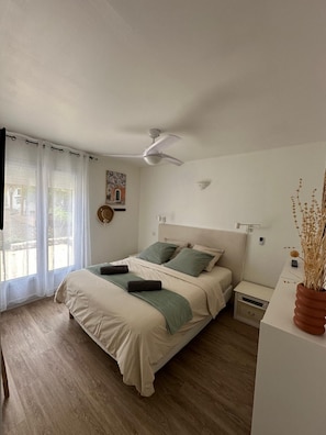 A bedroom with a bed, a ceiling fan, a nightstand, and a window with sheer curtains.