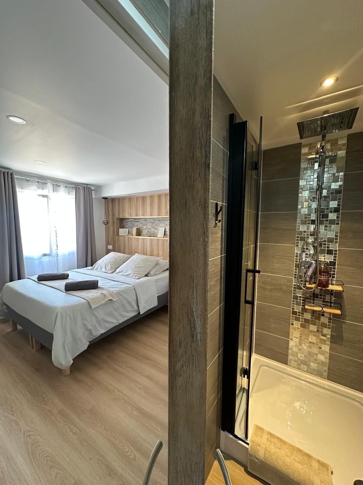 A modern hotel room with a large bed, a shower area with mosaic tiles, and a wooden floor.