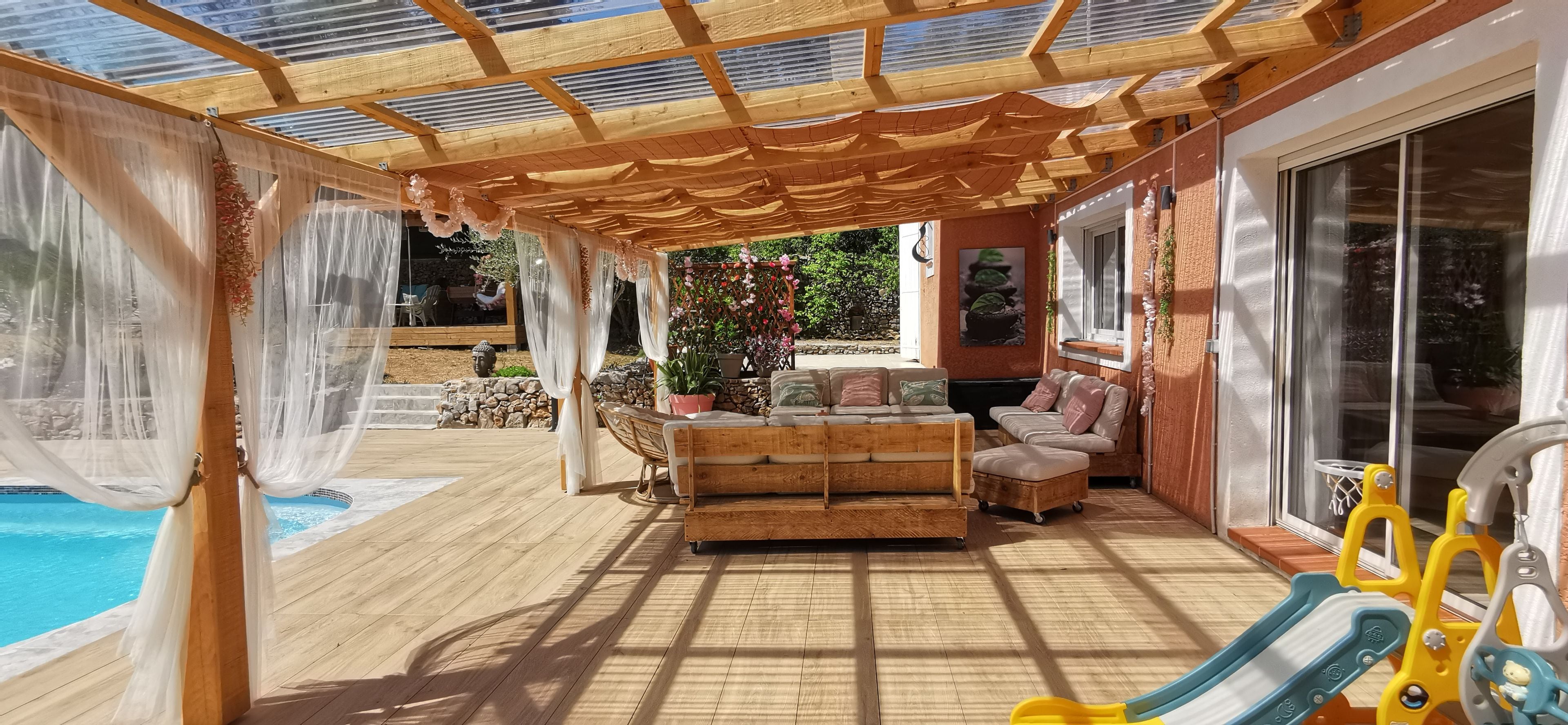 Ett takförsett utomhusområde med en pool, trämöbler och en solig atmosfär.