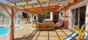 A covered outdoor area with a pool, wooden furniture, and a sunny ambiance.