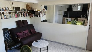 Een knus woongedeelte met een bank, een klein rond tafeltje en een groene stoel. Door een open deur is een keuken te zien.