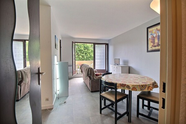 A room with a dining table, a sofa, and a small white cabinet. There is a door leading to another room and a window with a view of greenery outside.