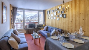 A cozy room with a sofa, dining table set for four, a TV, and a view of snowy mountains. - Wellmaintain sunny 3-room apartment in quiet residence with superb mountain view (Val-D'isère)