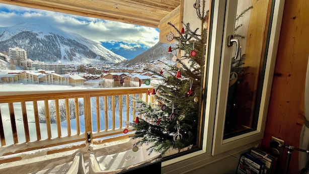 A balcony with a view of a snow-covered mountain town, a decorated Christmas tree, and a wooden railing. - Wellmaintain sunny 3-room apartment in quiet residence with superb mountain view (Val-D'isère)