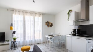 Moderni keittiö, jossa on valkoiset kaapit, musta liesi ja kahvinkeitin. Olohuoneessa on ruokapöytä tuoleineen ja televisio.