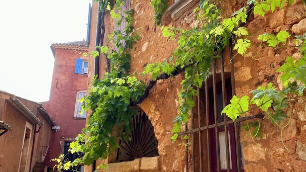 Un vicolo stretto con edifici in pietra rustica, uno con una porta in legno e un cancello in metallo, e un altro con una finestra e un arco in pietra.