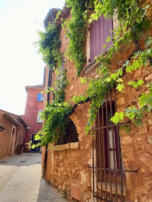 Une rue étroite bordée de bâtiments rustiques en pierre, dont l’un est doté d’une porte en bois et d’un portail en métal, et l’autre, d’une fenêtre et d’une arche en pierre.