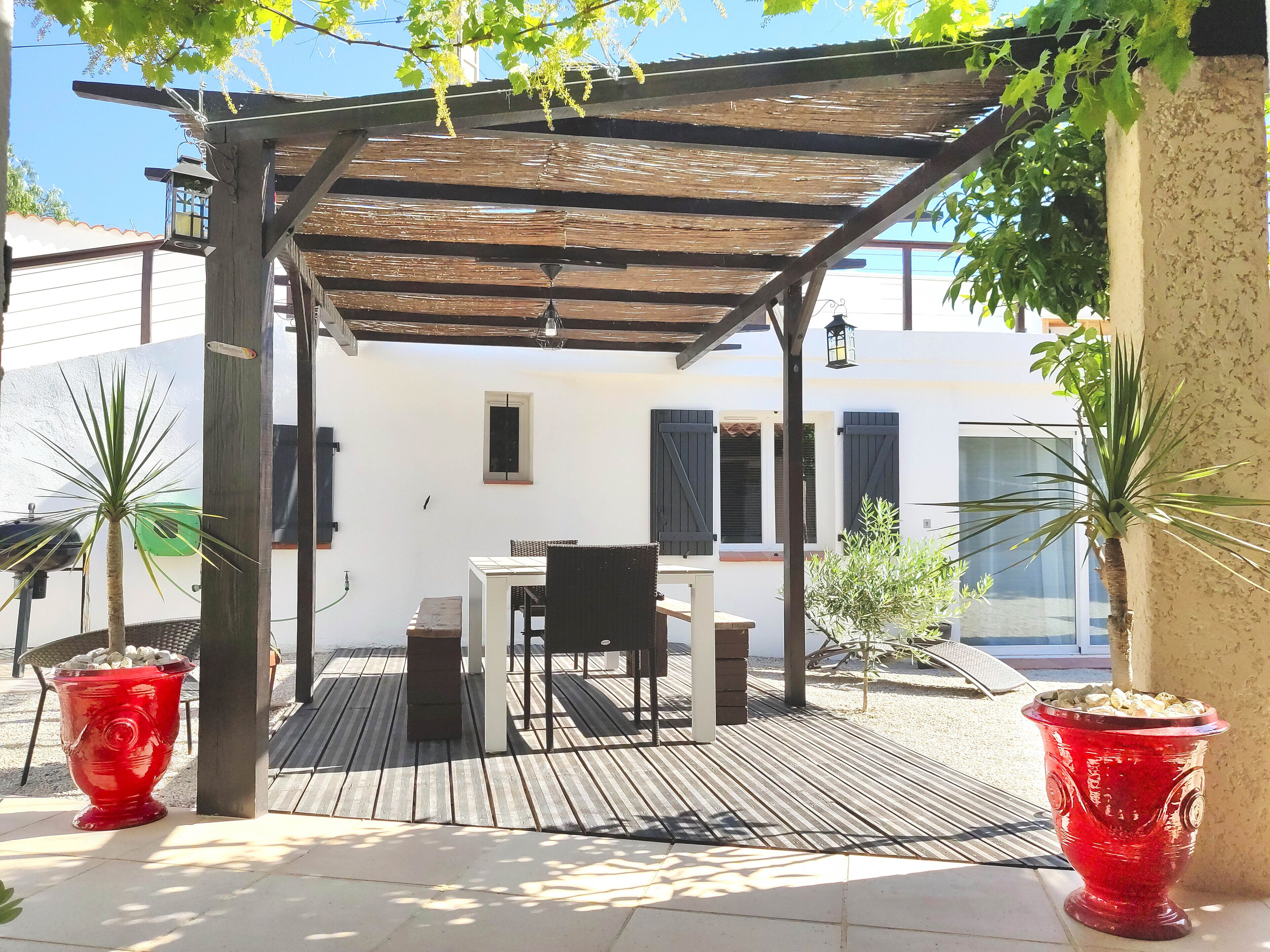 A covered patio with a table and chairs, surrounded by potted plants and a white building in the background.