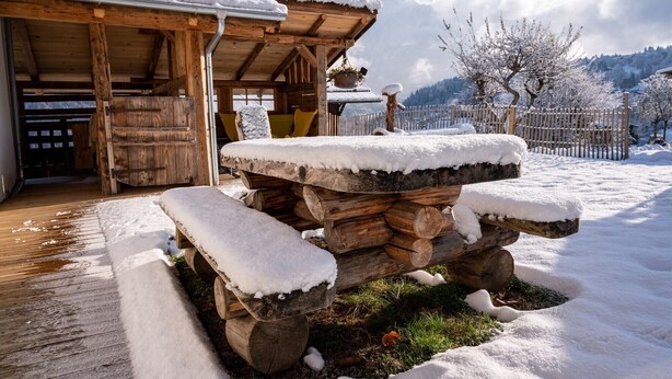 Eine hölzerne Außenbereich-Sitzgruppe mit Schneebedeckten Bänken, einer Holz-Pergola und Blick auf verschneite Bäume und Berge im Hintergrund.