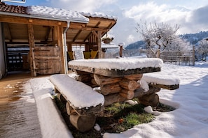 A wooden outdoor seating area with benches covered in snow, a wooden pergola, and a view of snow-covered trees and mountains in the background.