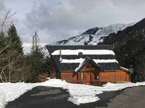 Una cabaña de madera con techo inclinado, rodeada de montañas y árboles cubiertos de nieve.