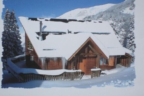 A wooden cabin with a snow-covered roof, surrounded by snow-covered trees and mountains.