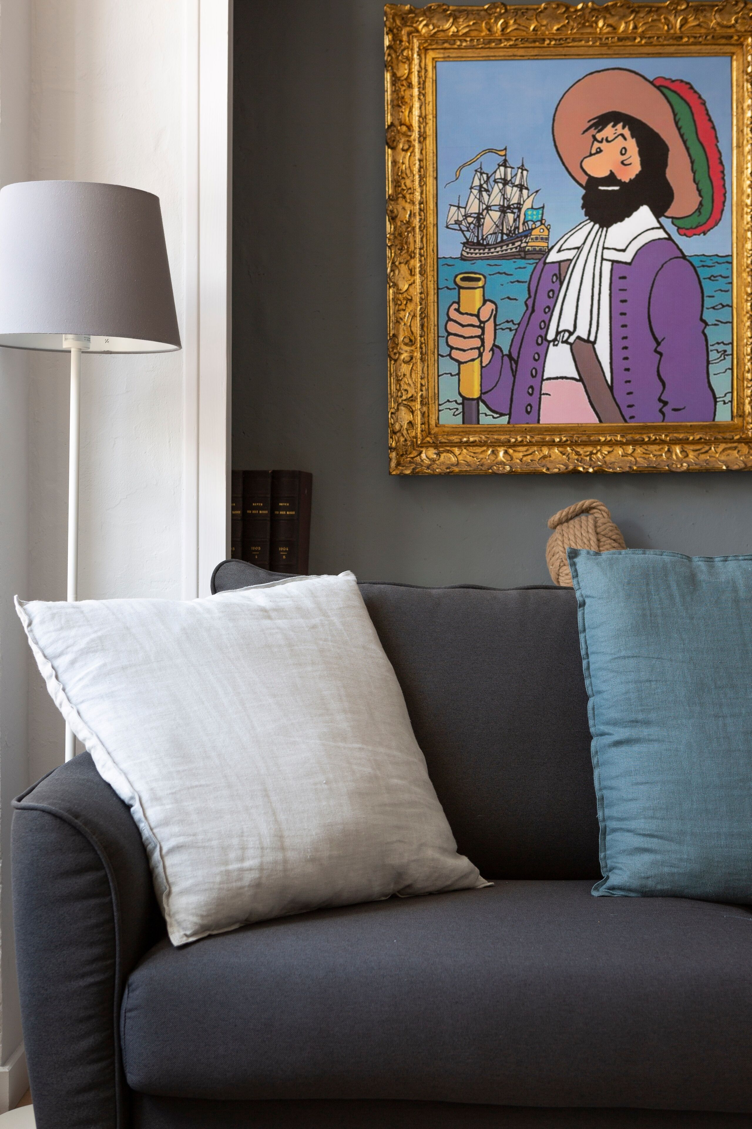 A dark-colored sofa with three pillows, a framed artwork depicting a historical figure, and a floor lamp.