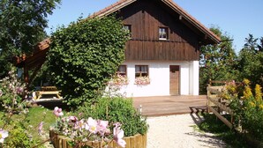 Una casa in legno con facciata bianca, un terrazzo in legno e un giardino con piante fiorite.