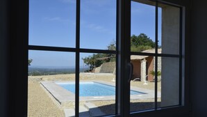 A view through a window of a swimming pool, stone patio, and a building with a stone facade. - Large Mas with heated pool for 10 people, 3 Bathrooms panomaric view (Cairanne)