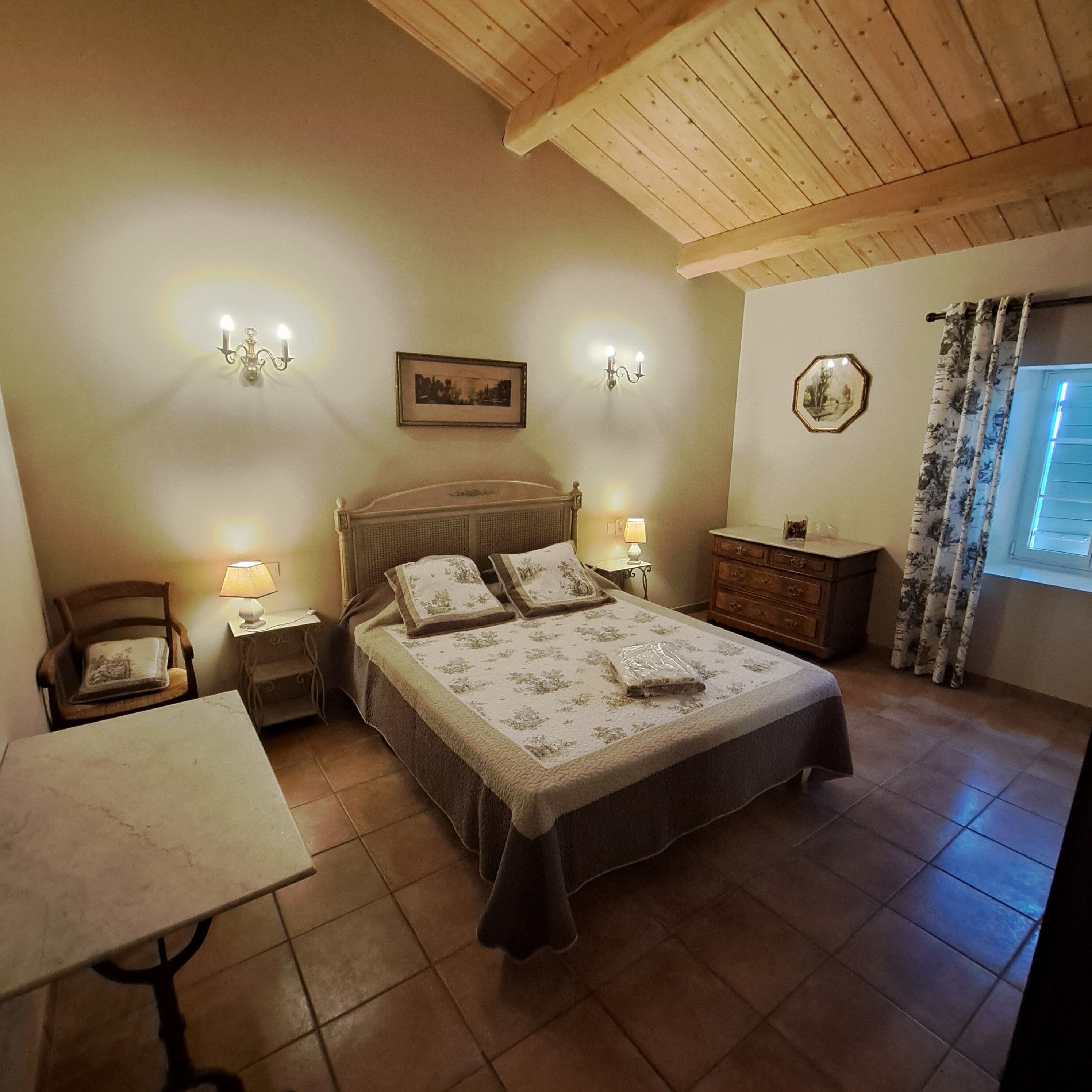 A bedroom with a wooden ceiling, a bed with a floral bedspread, two wall-mounted lamps, a nightstand, a chair, a dresser, and a window with curtains.