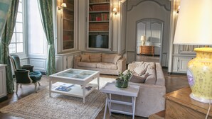 A classic living room with a glass coffee table, a beige sofa, a green armchair, and a bookshelf.
