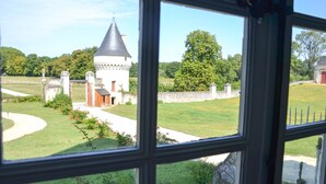 A view through a window of a castle-like structure with a tower, surrounded by a well-maintained lawn and a stone wall.